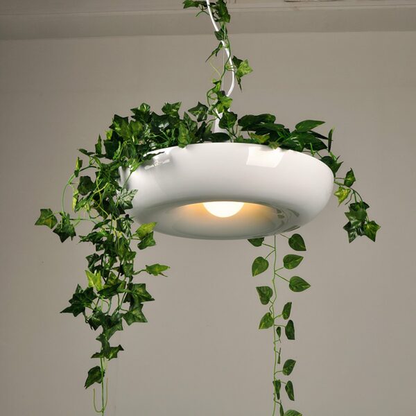 Suspended light, with white donuts shade, in which there is a drooping green plant. In the background, a white wall.