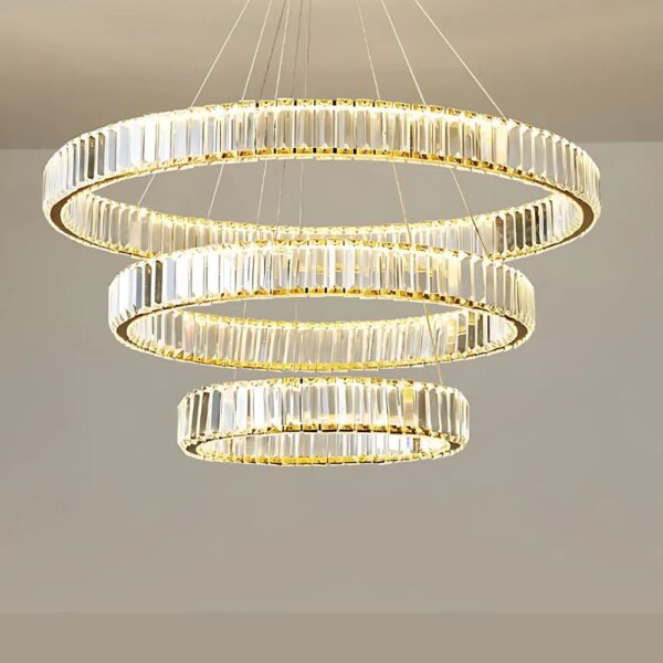 Large circular LED chandelier in gold chrome crystal, lit and installed in a living room, above a white sofa.