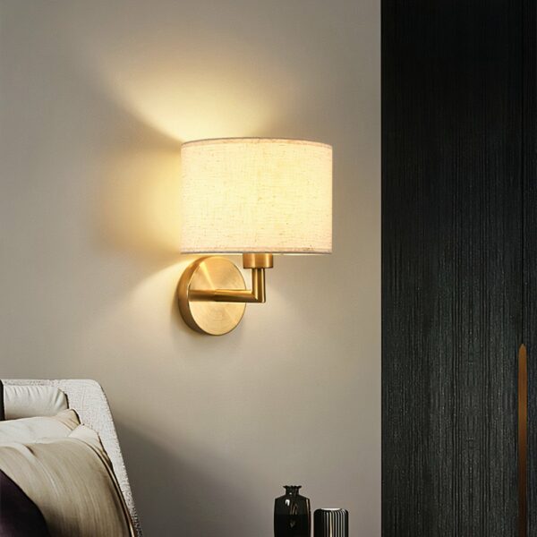 LED fabric wall light, with gold-colored metal bracket, light on, next to a bed.