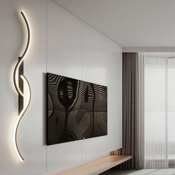 Photo of a large white wall on which hangs a large TV screen, to its left a 2-S-shaped designer wall light, one at the top, the other at the bottom, intertwined on a black rectangular base. A window can be seen in the background on the right.