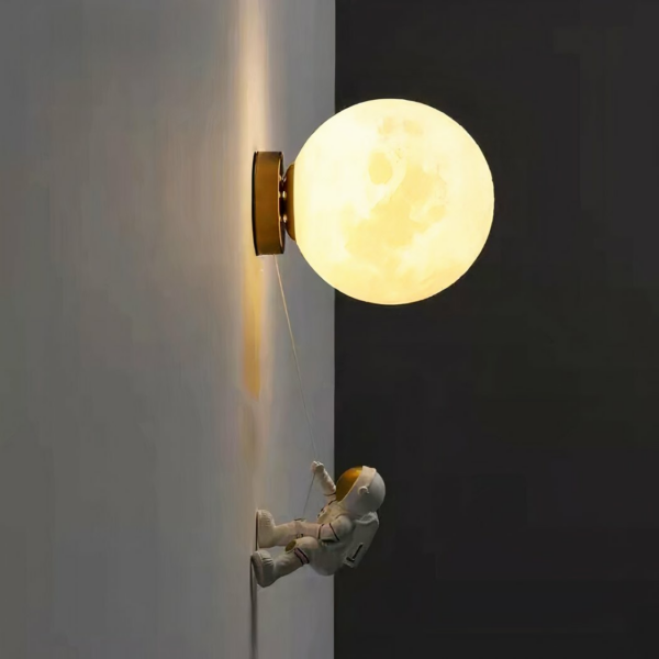 Wall-mounted sconce in the shape of a ball representing the moon, with a rope attached. At the end of the rope, an astronaut figure leans against the wall to climb to the moon. With the lights on, the astronaut stands on a white wall in a pitch-black room.