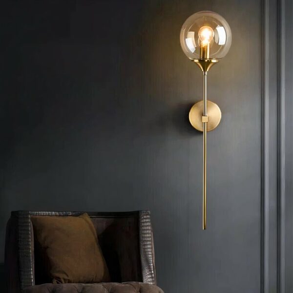 A sleek, modern wall sconce featuring a slim, gold-finish stem on which is a glass ball with a light bulb lit inside. The rod is attached to the wall by a round gold base. On a black wall. Next to it is an armchair, with only the brown backrest visible.