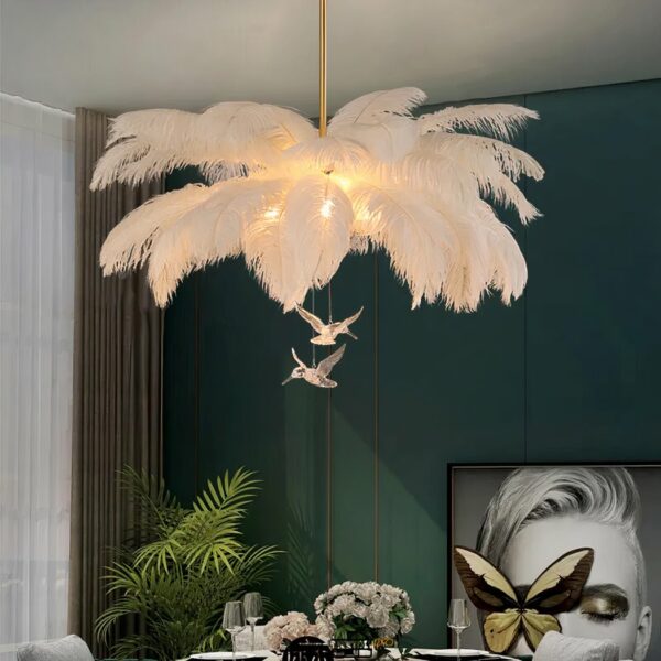 Chandelier with large white feathers and two transparent bird figures. Hanging. The bulbs are lit and camouflaged in the feathers. A dark green wall can be seen in the background, along with a painting and a green plant.