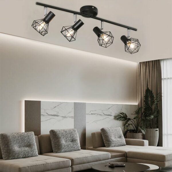 Black industrial-style ceiling chandelier with 4 industrial spotlight-style bulbs lined up. In a living room with a large beige corner sofa with 3 rectangular cushions. A green plant in the corner to the right of the sofa and a brown curtain behind it.