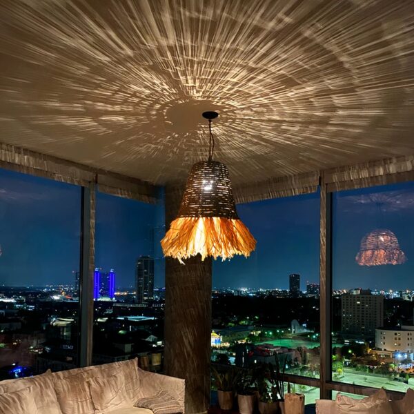 Hand-woven straw and raffia chandelier, hung from the ceiling and lit, in a living-room-style room. Illuminates the ceiling with the play of shadows created by the weaving of raffia and straw.