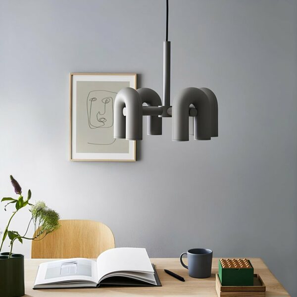 Grey chandelier with 4 upside-down U-shaped elements above a beige desk with an open book, a cup , a plant on the left, a beige chair. Behind a light gray wall with a picture frame.