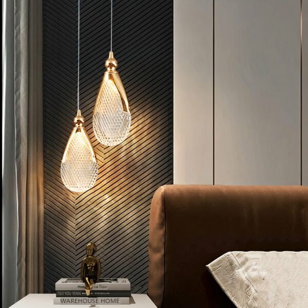 Suspended chandelier with 2 acrylic drops held by a gilded metal structure and fine wire. Lit and suspended above a bedside table with a Buddha, on the right a brown headboard, and a white cushion.