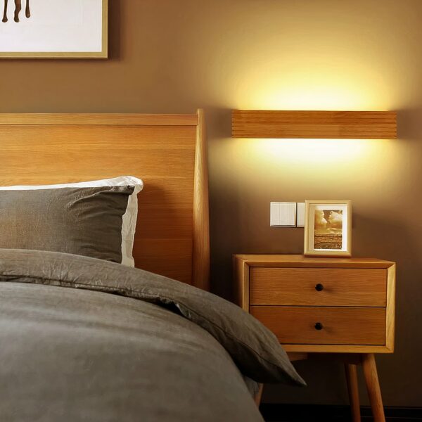 Wooden design staircase wall light hung in a bedroom and lit, next to the bed, on a taupe wall.