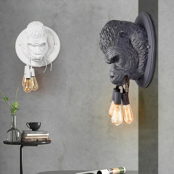 Two round wall sconces with the head of a 3D gorilla holding 3 strings in its mouth, with a light bulb hanging from each. On the grey wall opposite, the sconce is white, on the grey wall to the right, it's black. Under the white one, a round black table with a single slender leg. Above, a transparent vase with foliage, books and a mug on top of the stack of books.