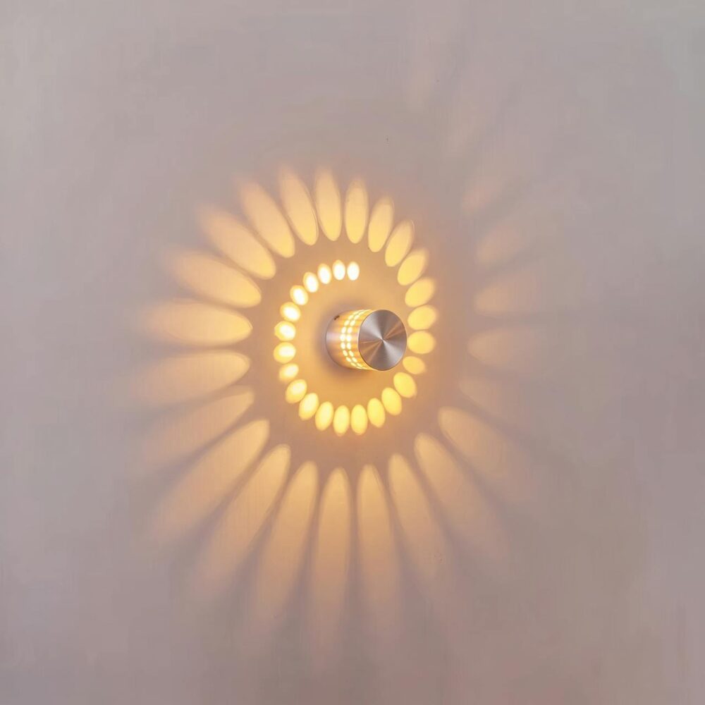 Round LED wall light in silver aluminium, switched on, casting a spiral of warm light on a white wall