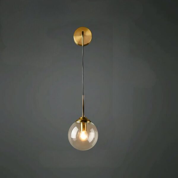 A modern wall sconce featuring a round gold base from which a slender gold stem extends downwards, at the end of which is a glass ball with a light bulb lit inside. On a black wall.