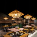 Lustre paille naturelle style paillote exotique accrochée au mur et allumée, au dessus de tables de ce qui semble etre un restaurant et dans une ambiance de lumière tamisée.