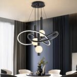 Black chandelier with black support fixed to the ceiling, from which cables descend to hold intertwined rounded structures that are luminous and black on the outside. In the center of these structures hang black tubes with a lighted ball. Fixed above a round white marble-style table with round black top, a vase on top and decorative objects. 6 armchairs around the table, to the left a bay window with a grey curtain and the wall opposite is duck-blue with vertical grooves.