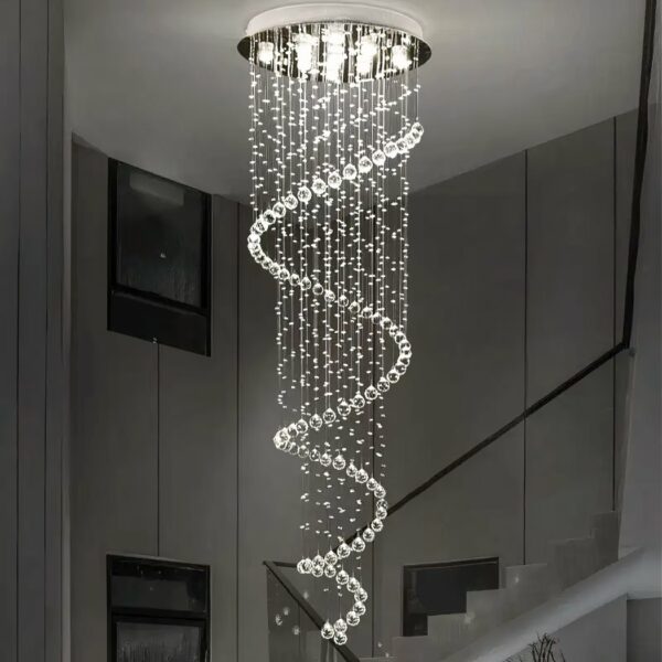 Ceiling chandelier in the shape of a spiral of teardrop suspensions. White light illuminated by several LEDs in the round silver ceiling bracket. Behind, a staircase with black railing and paintings on the wall.