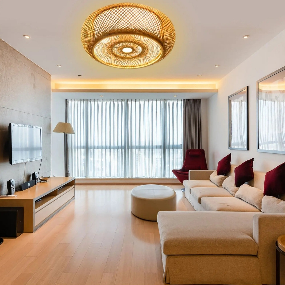 Lustre plafonnier rond en rotin allumé et fixé dans un salon. Sur la droite un grand canapé d'angle beige avec plusieurs coussins. En face, une grande fenêtre qui prend toute la largeur de la pièce. Sur la gauche, une télé au mur avec un meuble marron en dessous.