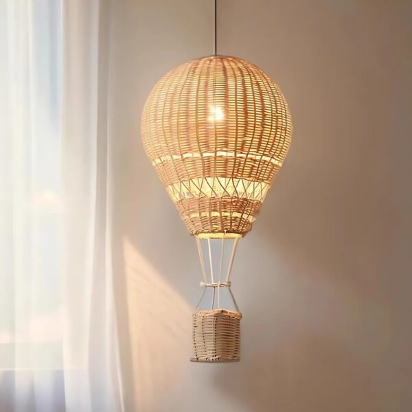 Lustre suspendu en forme de montgolfière avec une ampoule allumée au centre du ballon. Un mur beige derrière et une fenêtre avec voilage blanc à gauche.
