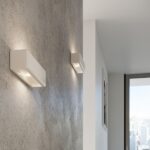 Two white rectangular wall lights on a textured grey wall near a window with a city view