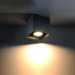 Black square ceiling spotlight mounted overhead casting a warm light beam