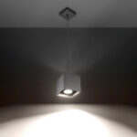 Grey cube pendant light hanging on a thin cable glowing on a dark ceiling
