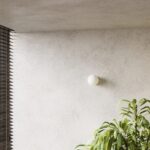Small round glass ball wall light on a textured wall beside tall green leaves