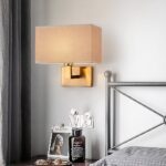 Brass wall light with a rectangular beige fabric drum shade beside a bed
