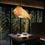 Woven bamboo fish-shaped pendant light glowing above a table near bamboo stalks