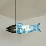 Single fish-shaped pendant lamp lit against a plain corner wall, showing the woven black head