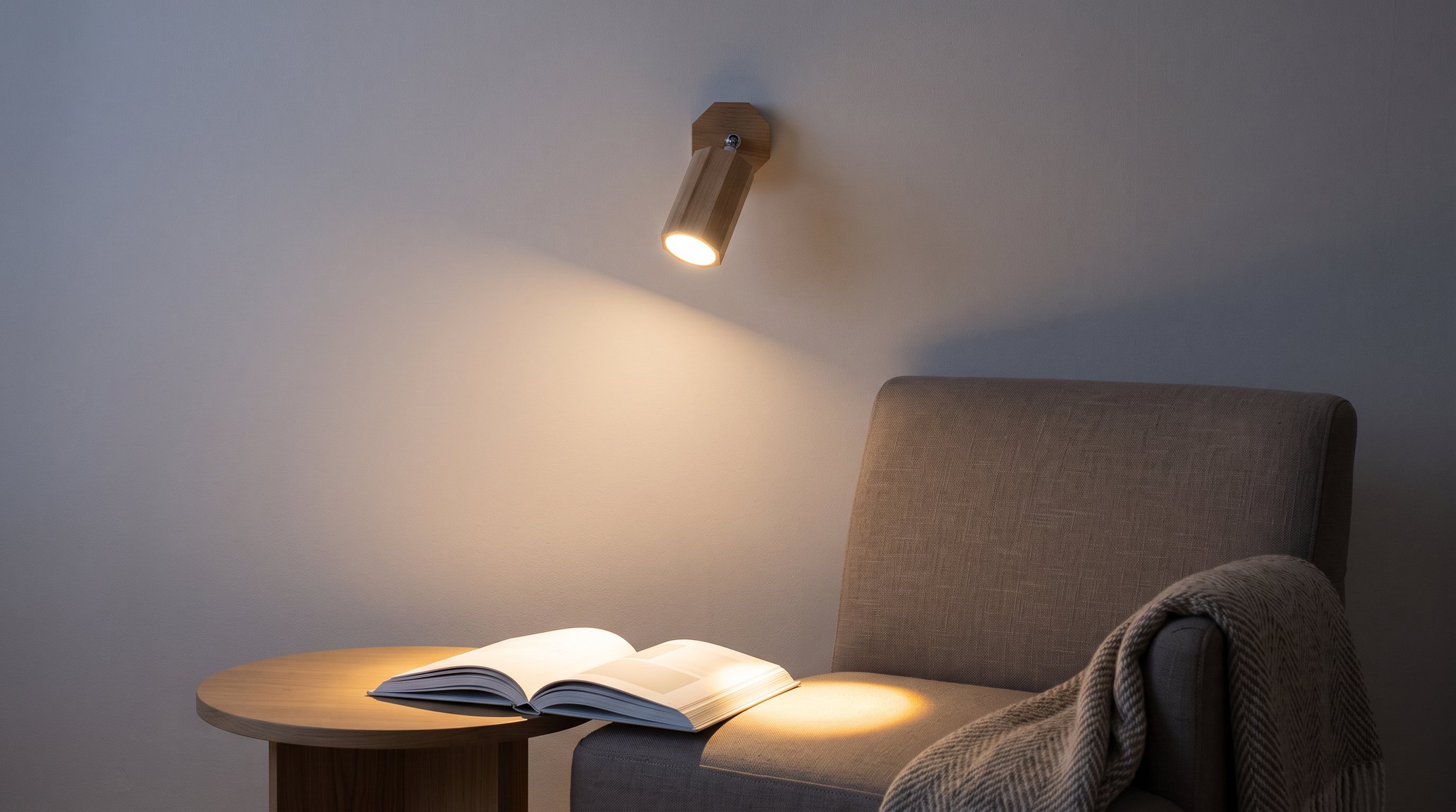 Reading corner in the evening, a natural oak wood spotlight wall light angled downwards, its warm beam forming a pool of light on the seat of a taupe linen armchair next to an open book on a round wooden side table