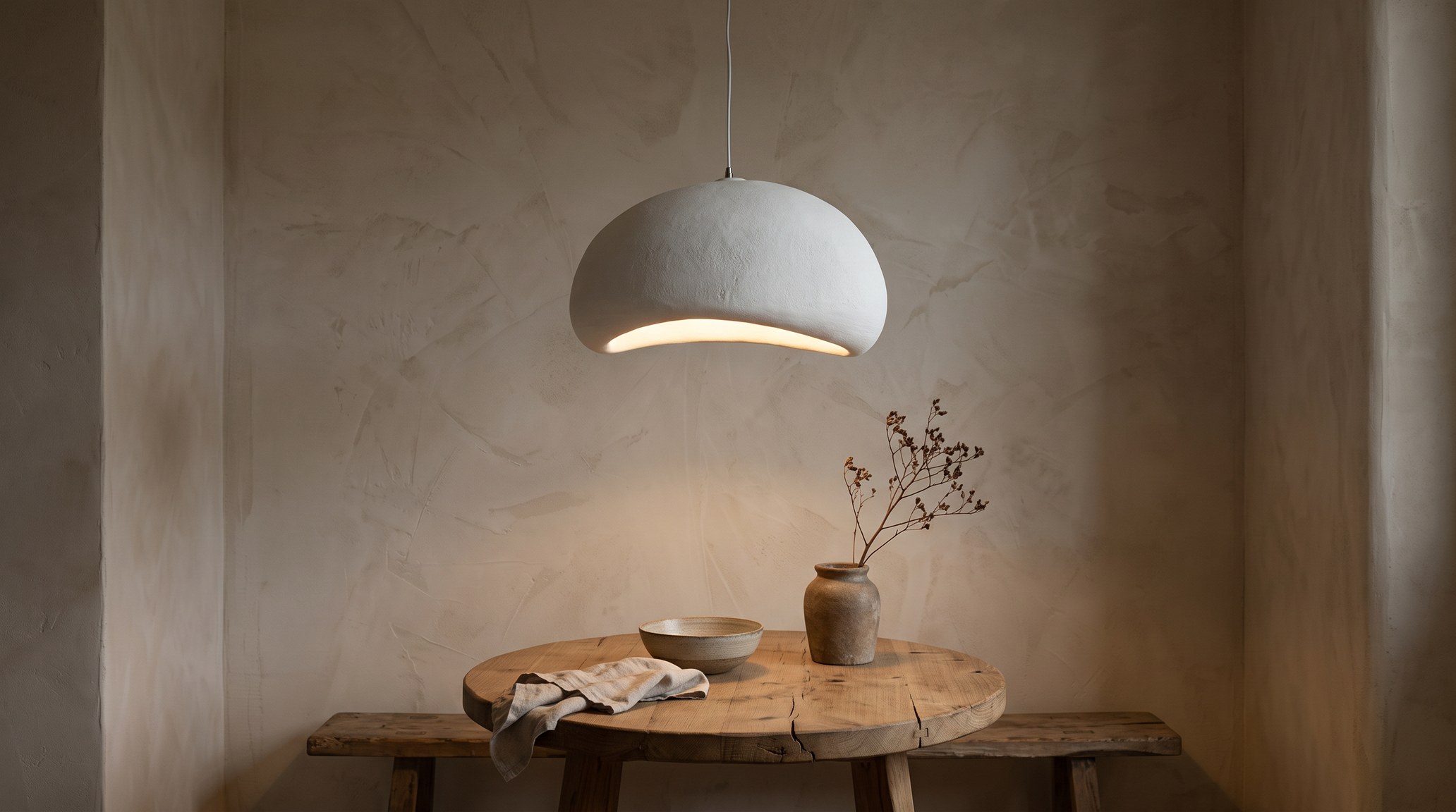 Wabi-sabi dining corner at dusk, a matt white pebble-shaped pendant lit in warm white above a round raw wooden table, a limewashed wall, a stoneware bowl and a dried branch